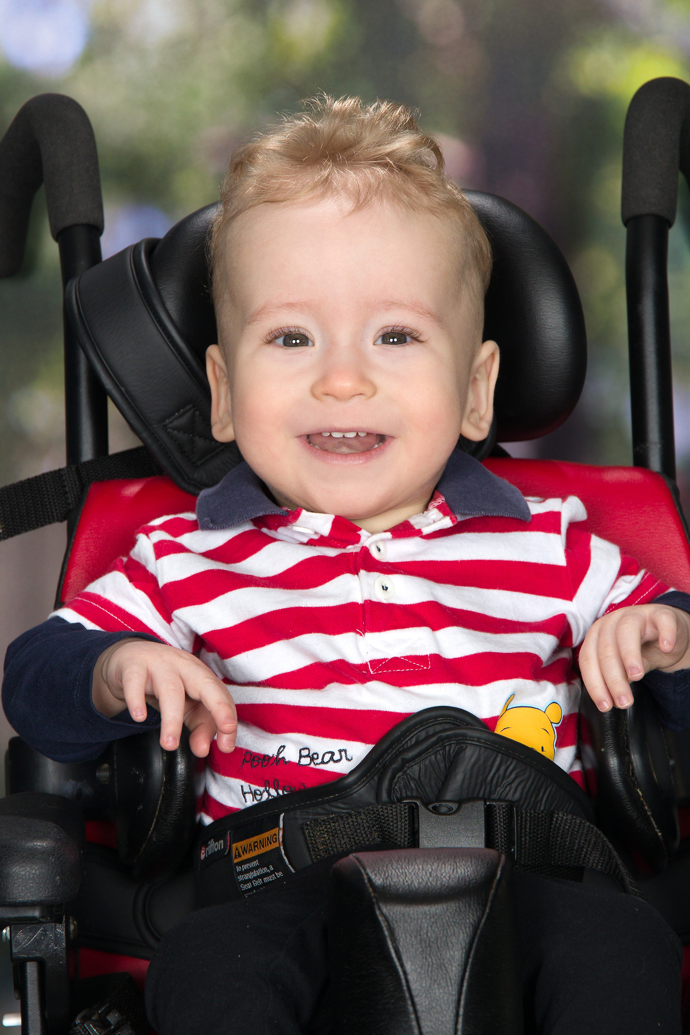 Adam at one year old, smiling for the camera, sitting in his wheelchair.