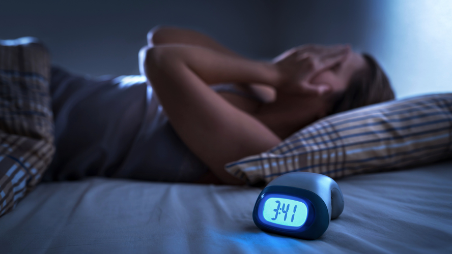 A woman lying awake in bed after having a nightmare. Here hands cover her face and a digital clock in the foreground shows 3:41am