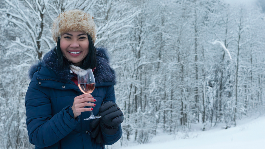 A woman holding a glass of wine while standing in snow in front of a snowy forest