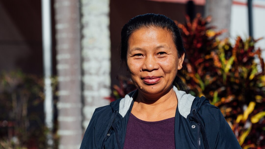 A portrait of a woman smiling at the camera. Her hair is pulled back and there are plants in the background.